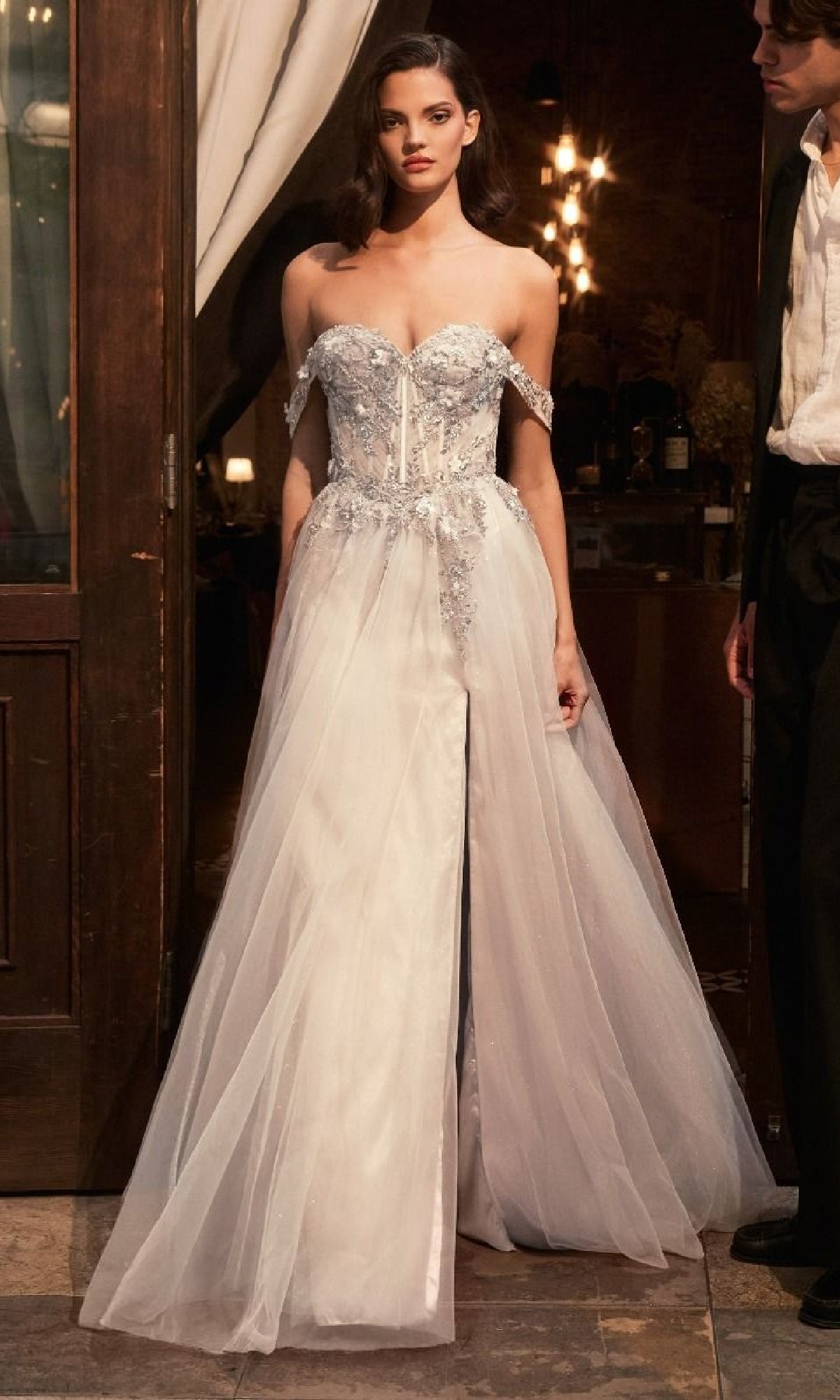 A woman wearing the Sheer-Corset White Bridal Gown: Ladivine KV1126W with an embellished sweetheart bodice and tulle skirt stands indoors beside a man in formal attire.